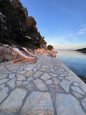 A stone-paved walkway with lounge chairs by a calm body of water, surrounded by rocky terrain and trees. - Apartments Rusulica (Marina)