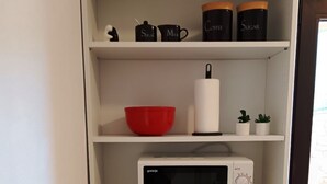 Una cocina con un microondas blanco, un cuenco rojo, un hervidor de acero inoxidable y dos pequeñas macetas con plantas en el estante.