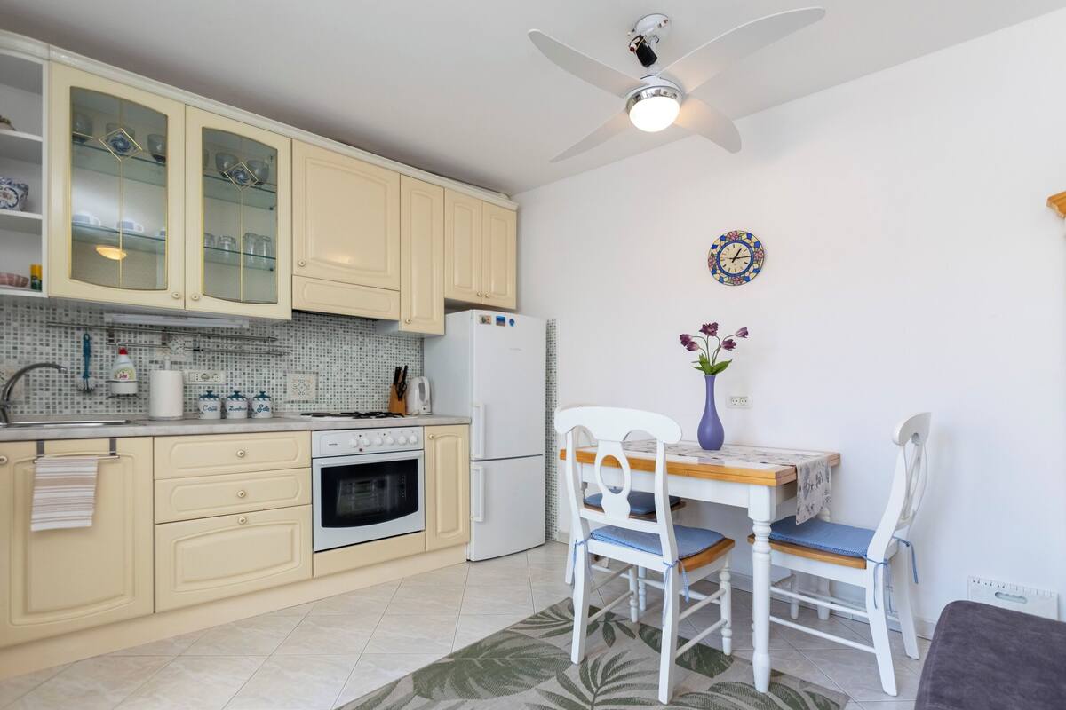 A kitchen with cream-colored cabinets, a white refrigerator, a stove, and a dining area with a table and chairs.