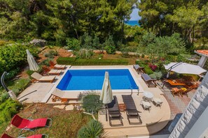 A rectangular swimming pool with lounge chairs and umbrellas, surrounded by lush greenery and a view of the sea. - House Sanda (Supetar)