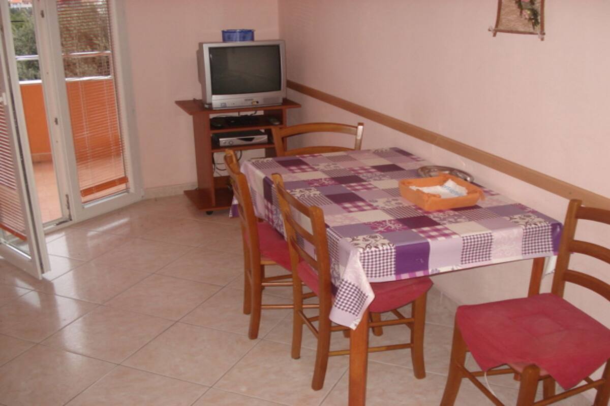 Un coin repas avec une table recouverte d’une nappe à carreaux, entourée de chaises en bois avec des coussins rouges. Un téléviseur est posé sur un meuble en bois, adossé au mur.