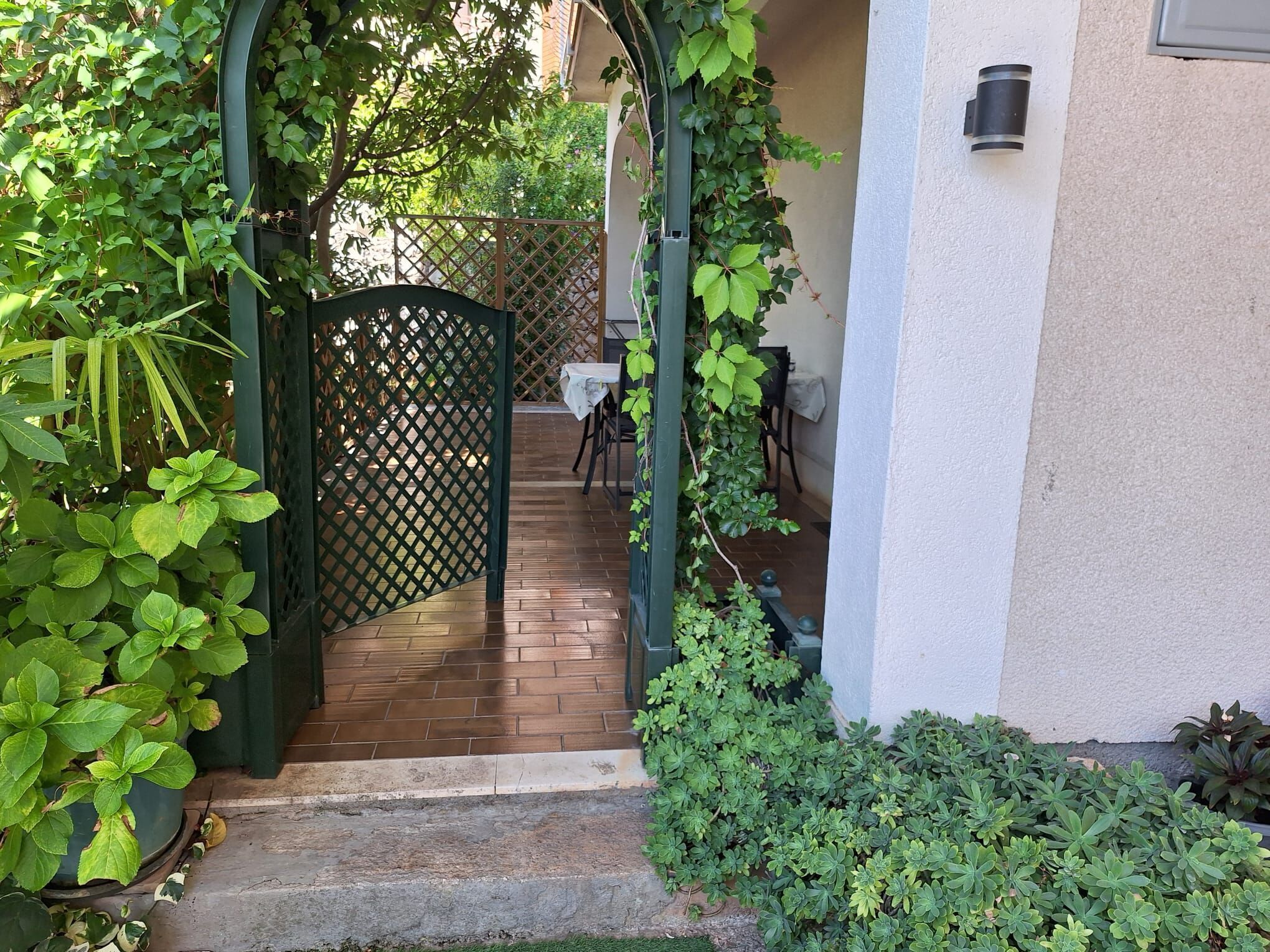 Un patio cubierto con suelo de madera, rodeado de follaje y un portón celosía.