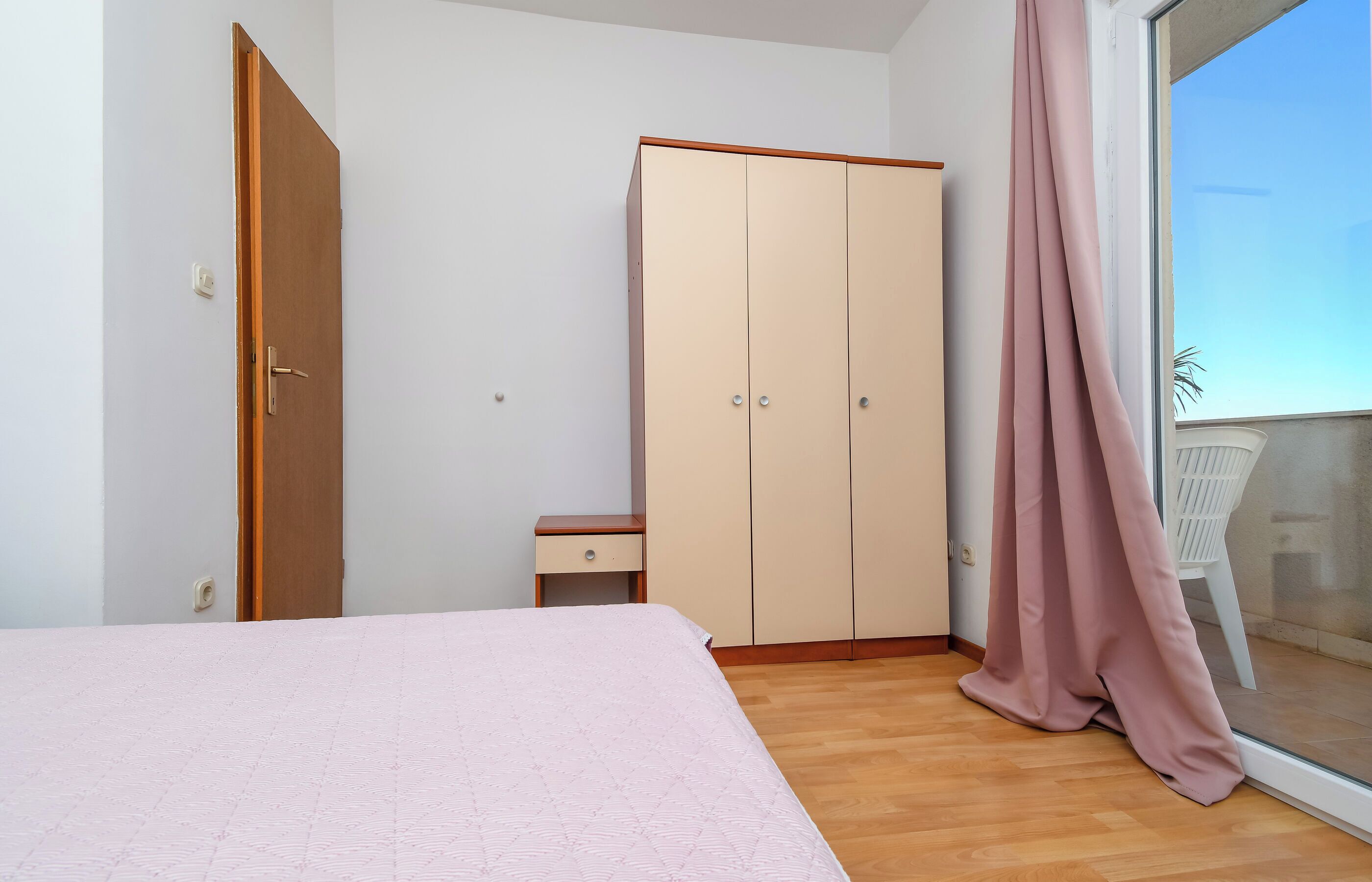 A bedroom with a bed, a wardrobe, a small bedside table, and a window with curtains.