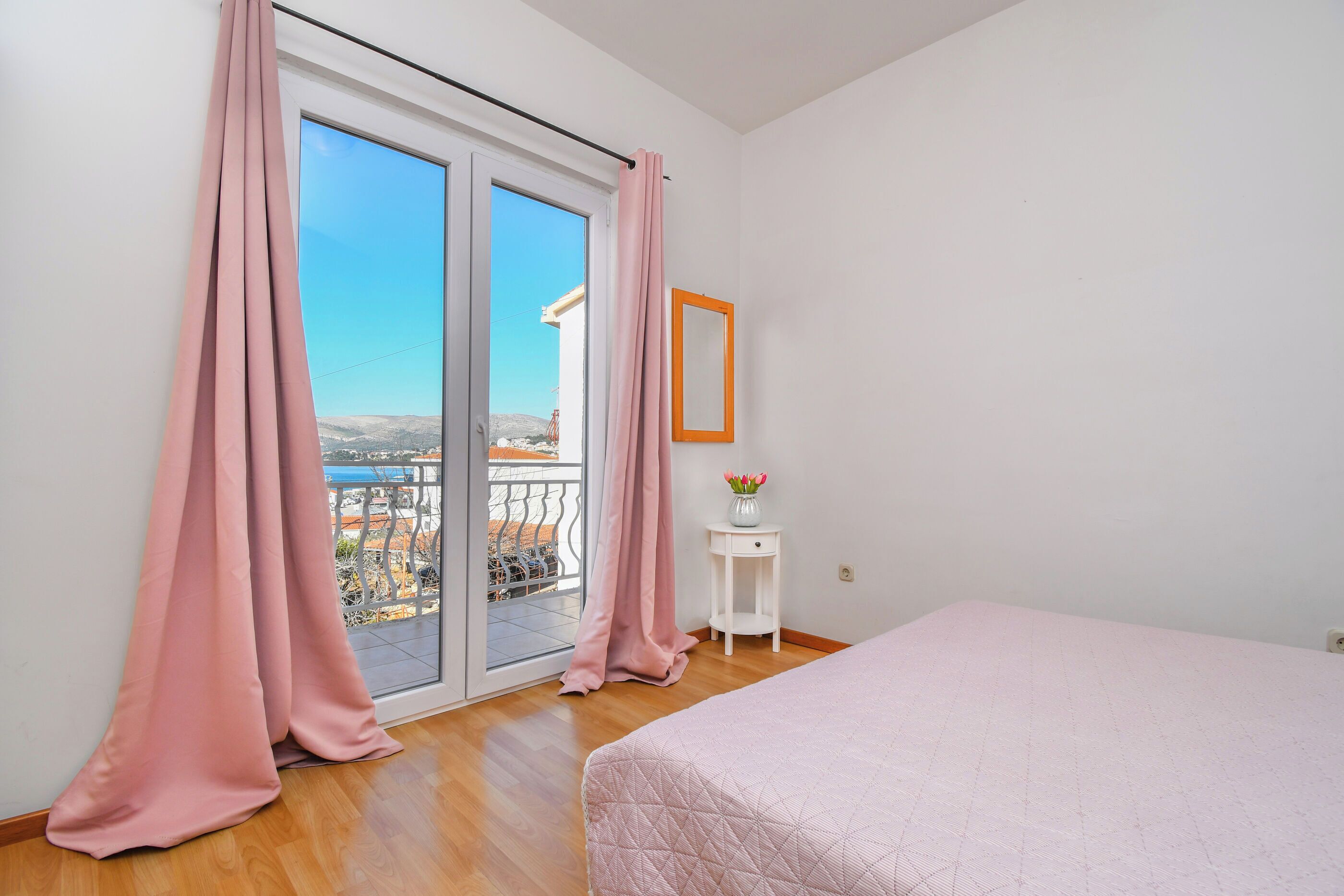 A bedroom with a balcony, a bed, a small white table with a vase, and a mirror on the wall.