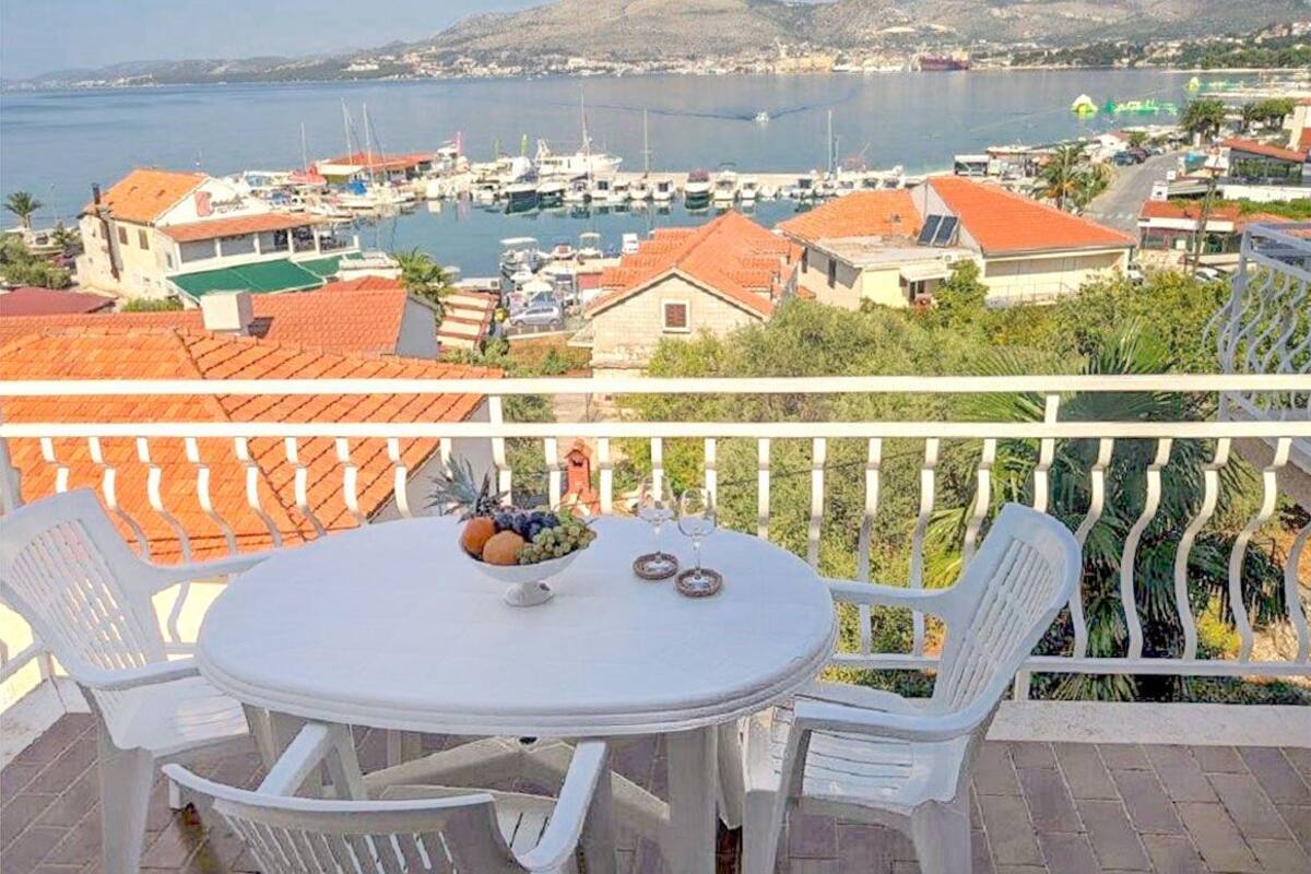 A balcony with a table and chairs overlooking a marina with boats and a mountainous landscape.