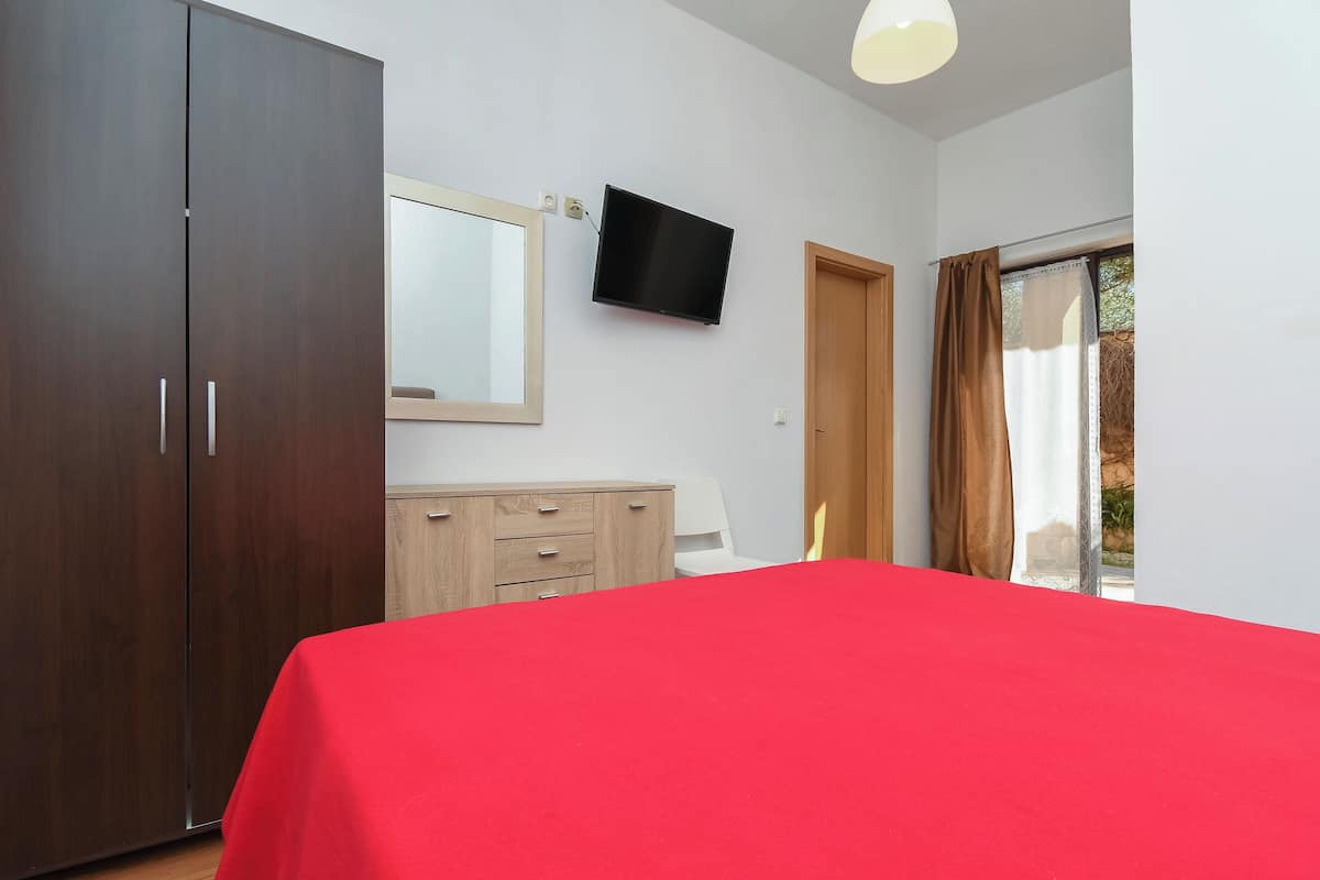 A bedroom with a large wardrobe, a bed with a red cover, a television on the wall, and a window with curtains.