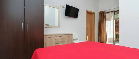 A bedroom with a large wardrobe, a bed with a red cover, a television on the wall, and a window with curtains.