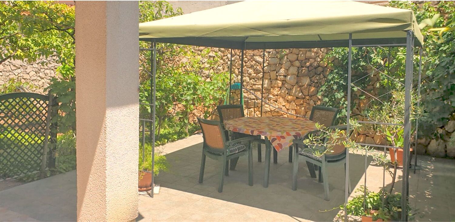 Un patio al aire libre con una mesa y sillas bajo una cubierta, rodeado por un muro de piedra y follaje.