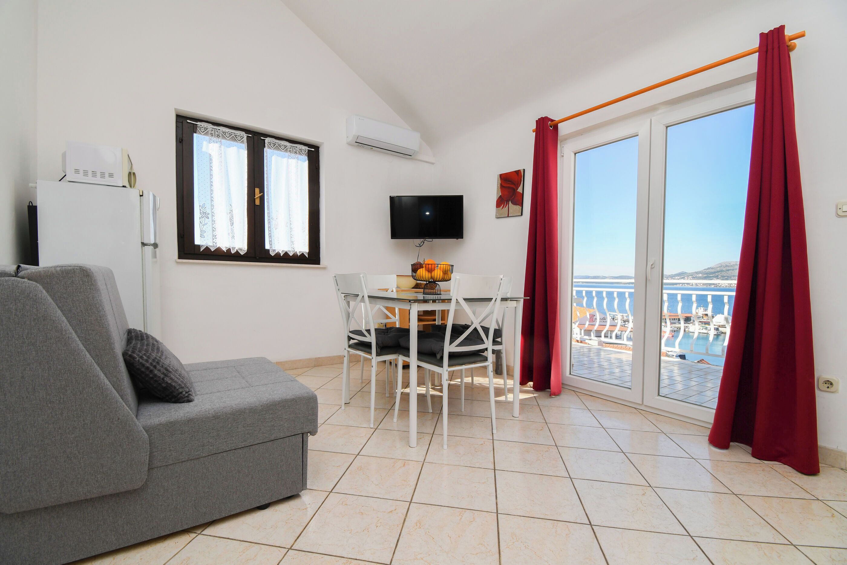 A living room with a grey sofa, a dining table with chairs, a TV, and a balcony with red curtains.