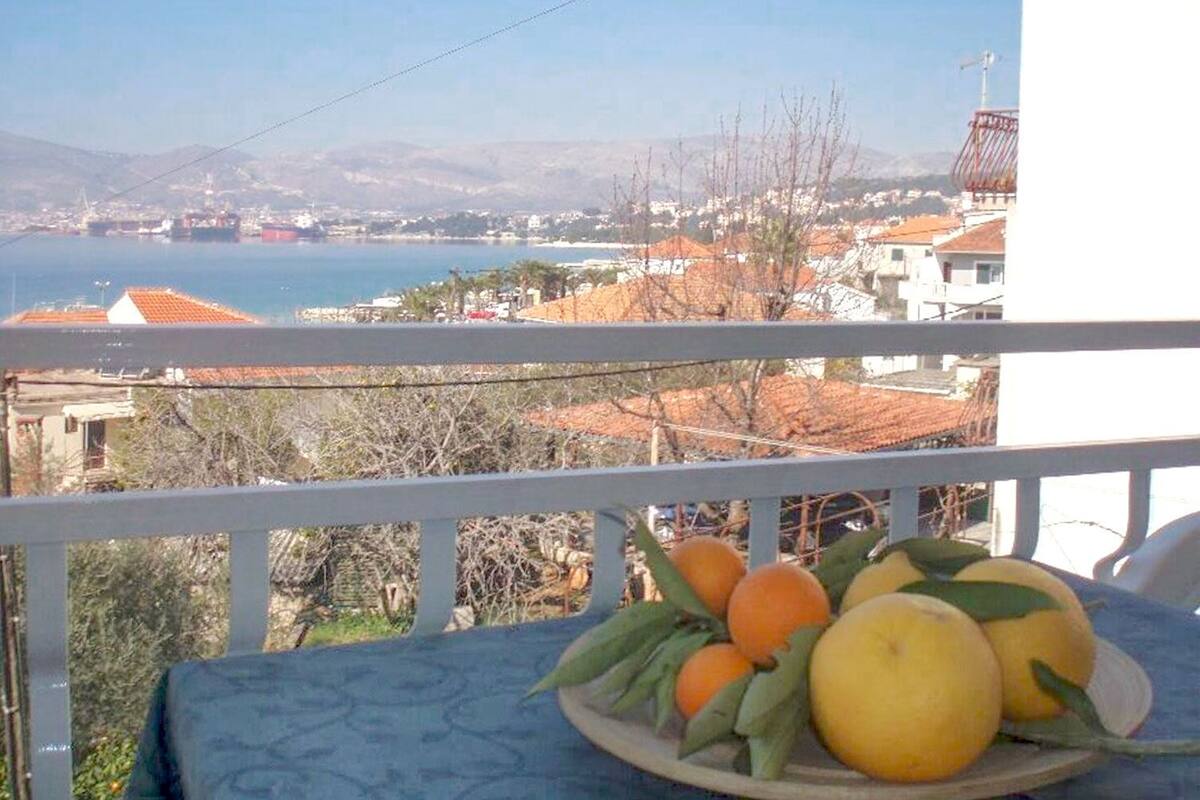 A balcony with a table holding oranges and lemons, overlooking a coastal town with buildings and a sea view.