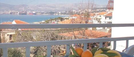 A balcony with a table holding oranges and lemons, overlooking a coastal town with buildings and a sea view.
