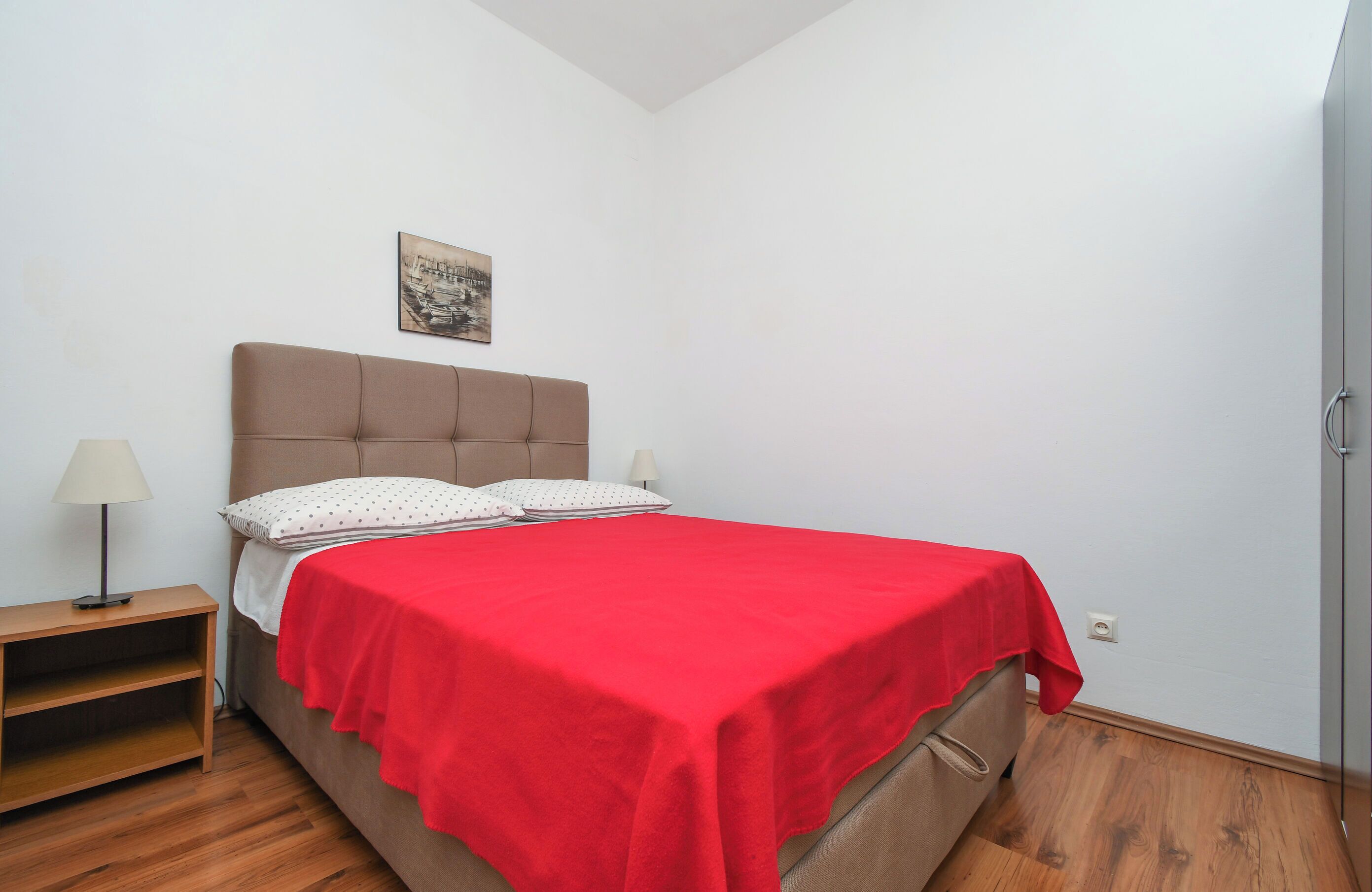Un dormitorio con cama, mesita de noche, lámpara, colcha roja y un cuadro en la pared.