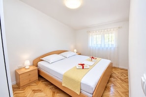 Una cama individual con cabecera de madera, dos lámparas de noche, una mesita de noche con cajón y una ventana con cortinas translúcidas.