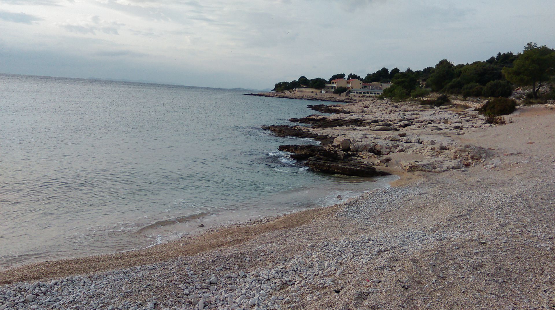 A pebble beach with a rocky shoreline, a building with a balcony overlooking the sea, and trees in the background.