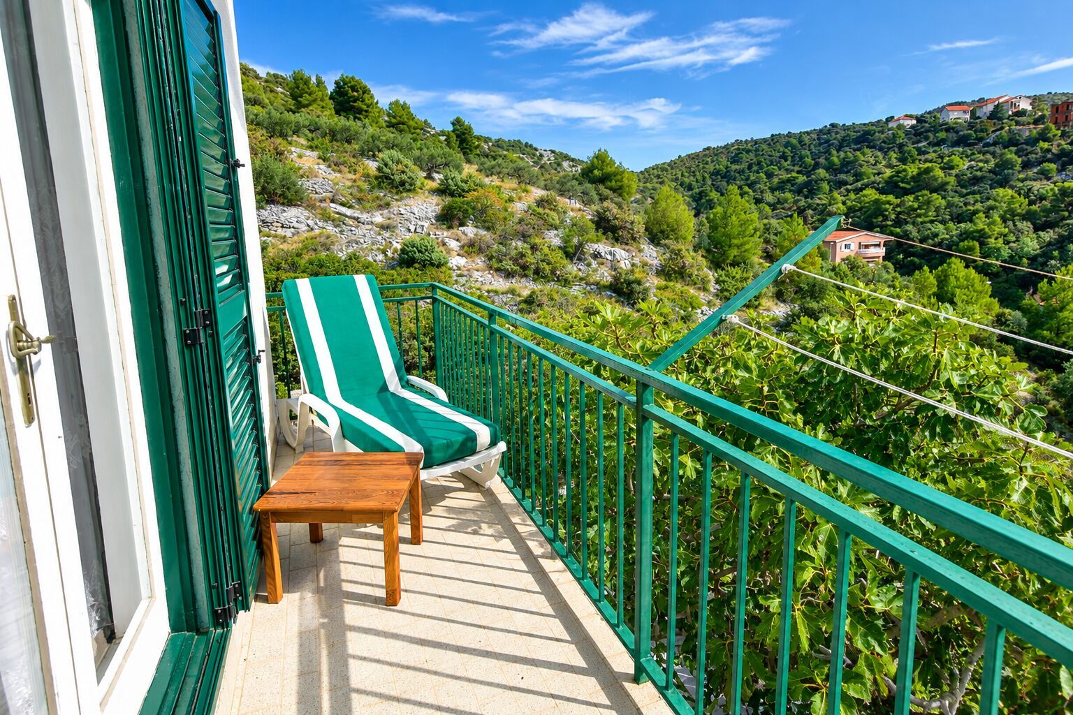 A balcony with two green-striped lounge chairs and a small wooden table, overlooking a hillside with vegetation and buildings.