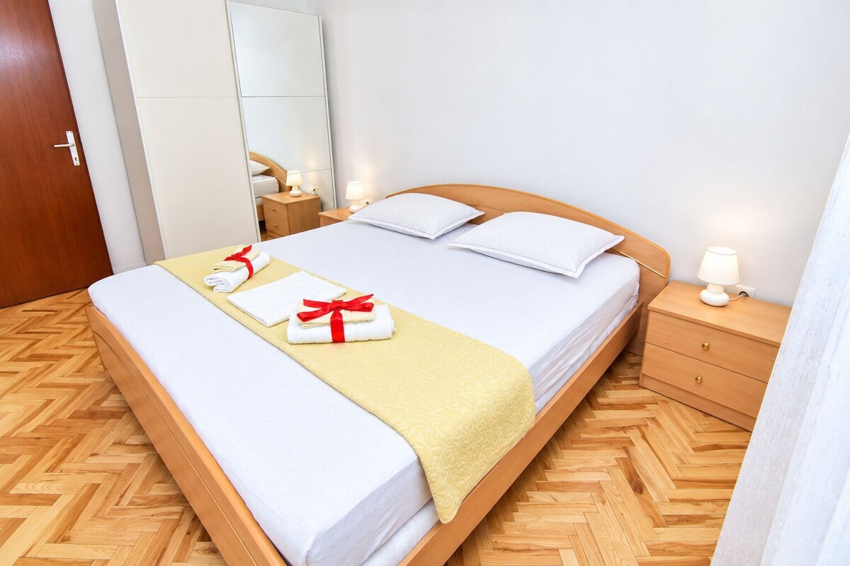 A neatly made bed with white linens and a yellow blanket, a wooden nightstand with a lamp, and a wooden floor with a herringbone pattern.