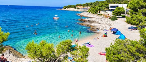 A coastal beach with clear waters, rocky shoreline, and a few buildings in the background.