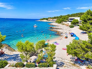 A coastal beach with clear waters, rocky shoreline, and a few buildings in the background. - Apartments Neva (Primosten)