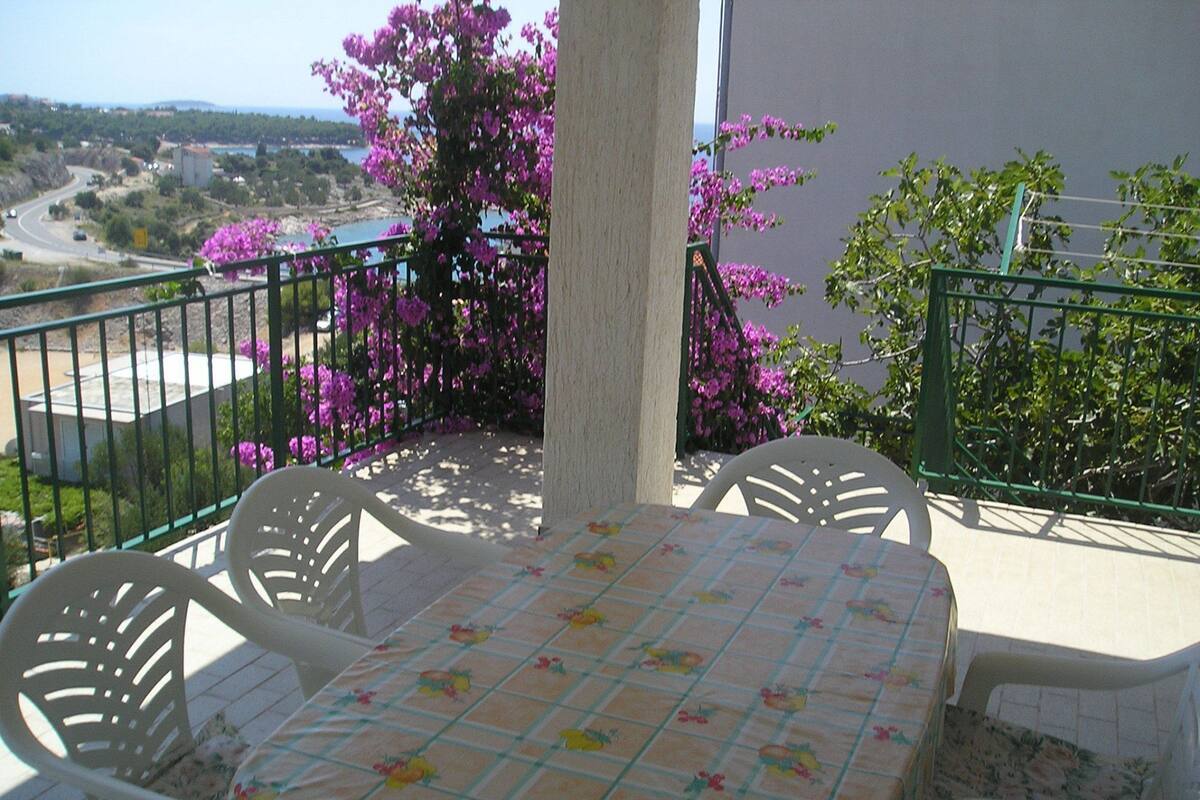 A balcony with a table and chairs, overlooking a coastal landscape with purple flowers and a view of the sea.