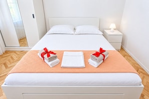 A hotel room with a bed, two towels with red ribbons, a nightstand with a lamp, and a wooden floor. - Apartments Neva (Primosten)