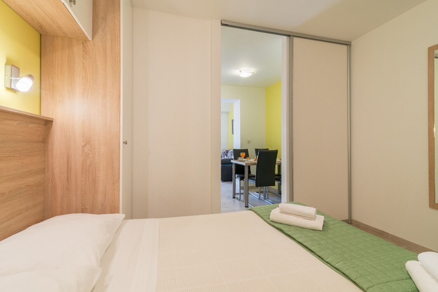 A hotel room with a bed, a green area rug, a dining area visible through a sliding door, and a mirror on the wall.