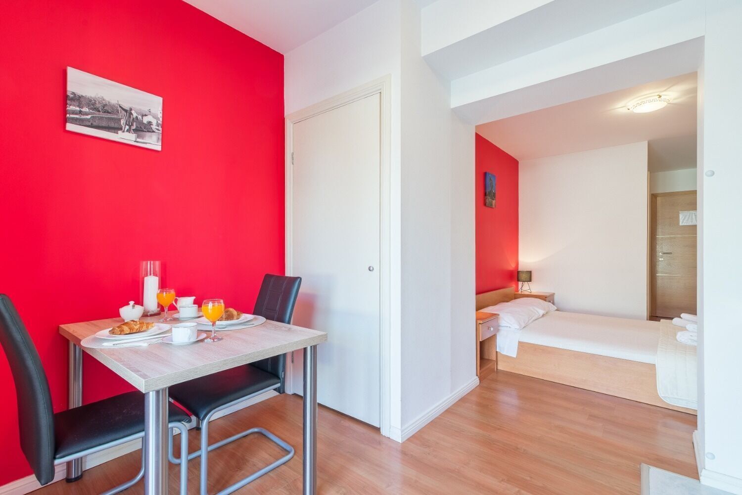 A compact hotel room with a dining area featuring a table set for breakfast, a bed with a wooden headboard, and a small bedside table with a lamp.
