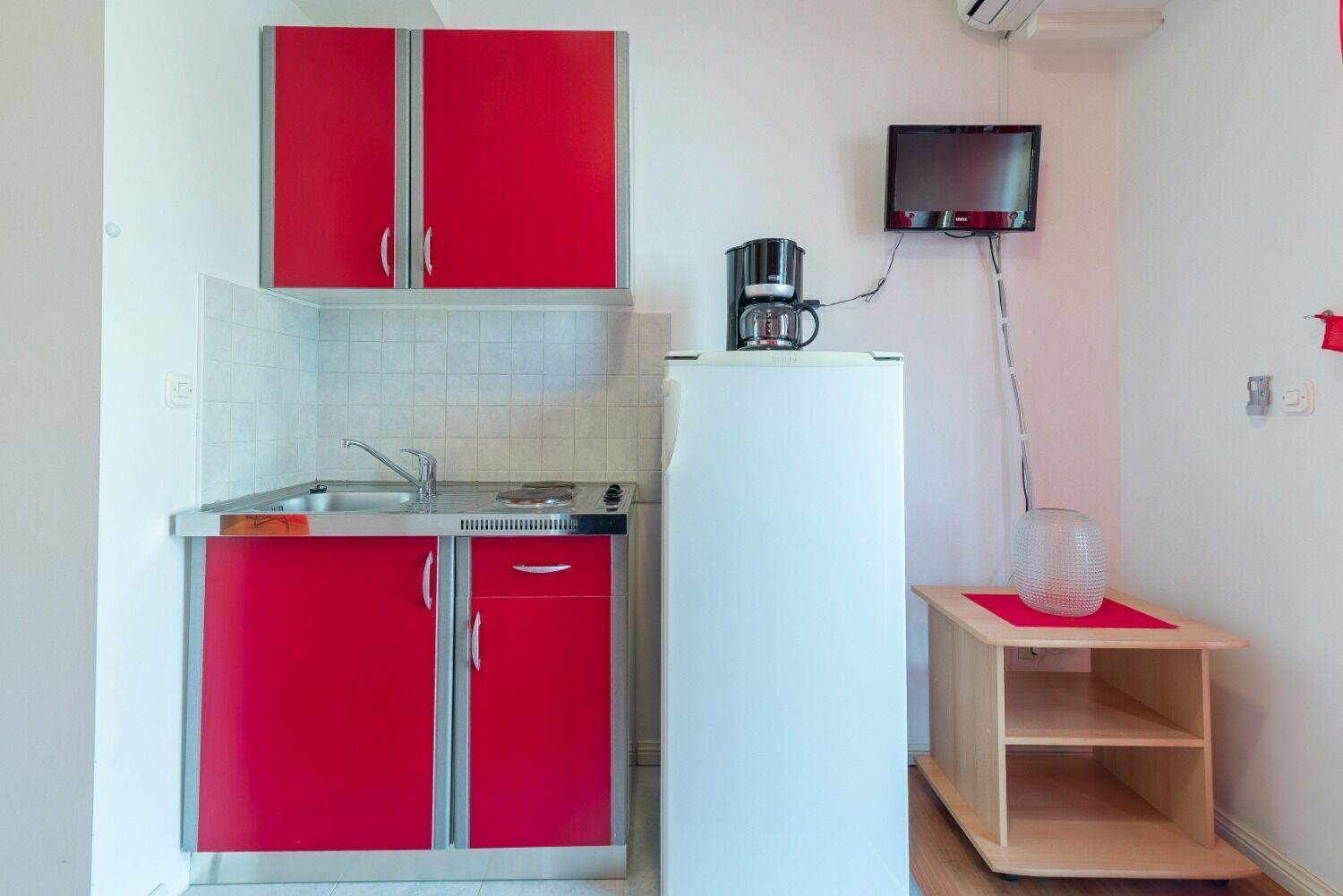 A compact kitchen with red cabinets, a white refrigerator, a coffee maker, and a small wooden table.
