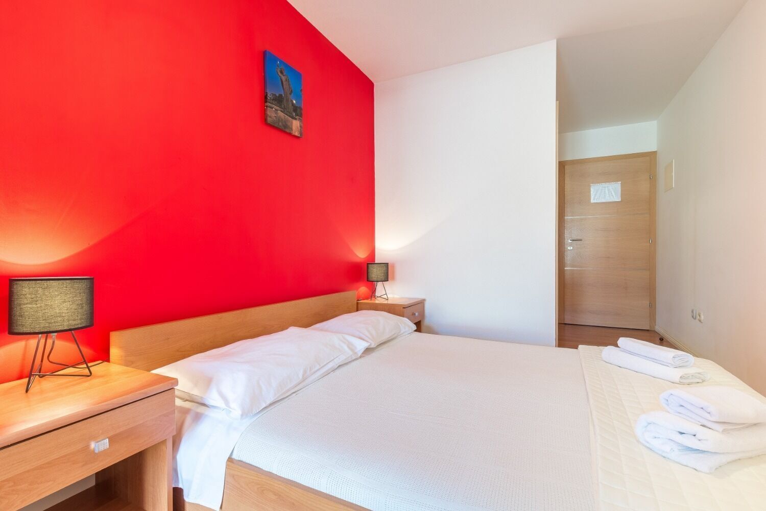 Habitación de hotel con una pared de acento roja, cama con ropa blanca, mesita de noche con lámpara y puerta de madera.