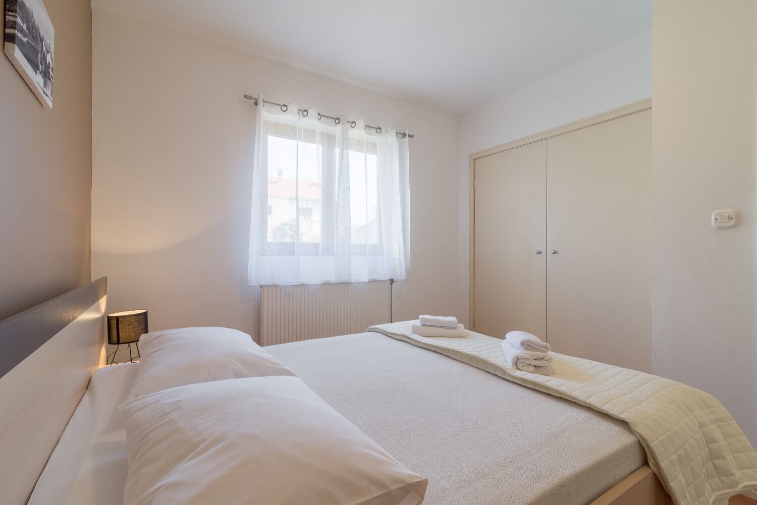 Ein Schlafzimmer mit Bett, Nachttischlampe, Fenster mit durchsichtigen Vorhängen, Heizkörper und Kleiderschrank.