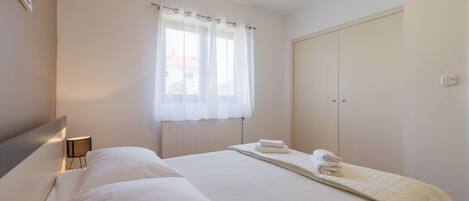 Ein Schlafzimmer mit Bett, Nachttischlampe, Fenster mit durchsichtigen Vorhängen, Heizkörper und Kleiderschrank.