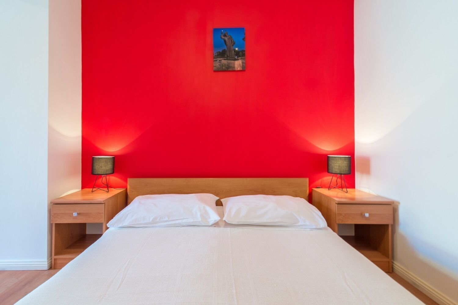 A hotel room with a red accent wall, a bed with white linens, two bedside tables with lamps, and a painting above the bed.