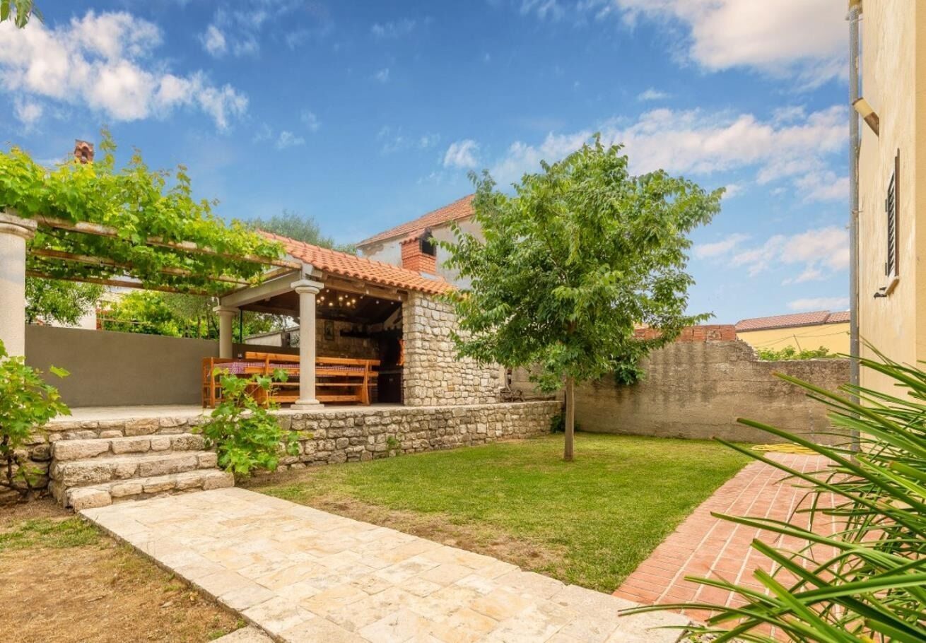 A stone building with a tiled roof, a stone staircase leading to a covered patio, and a grassy yard.