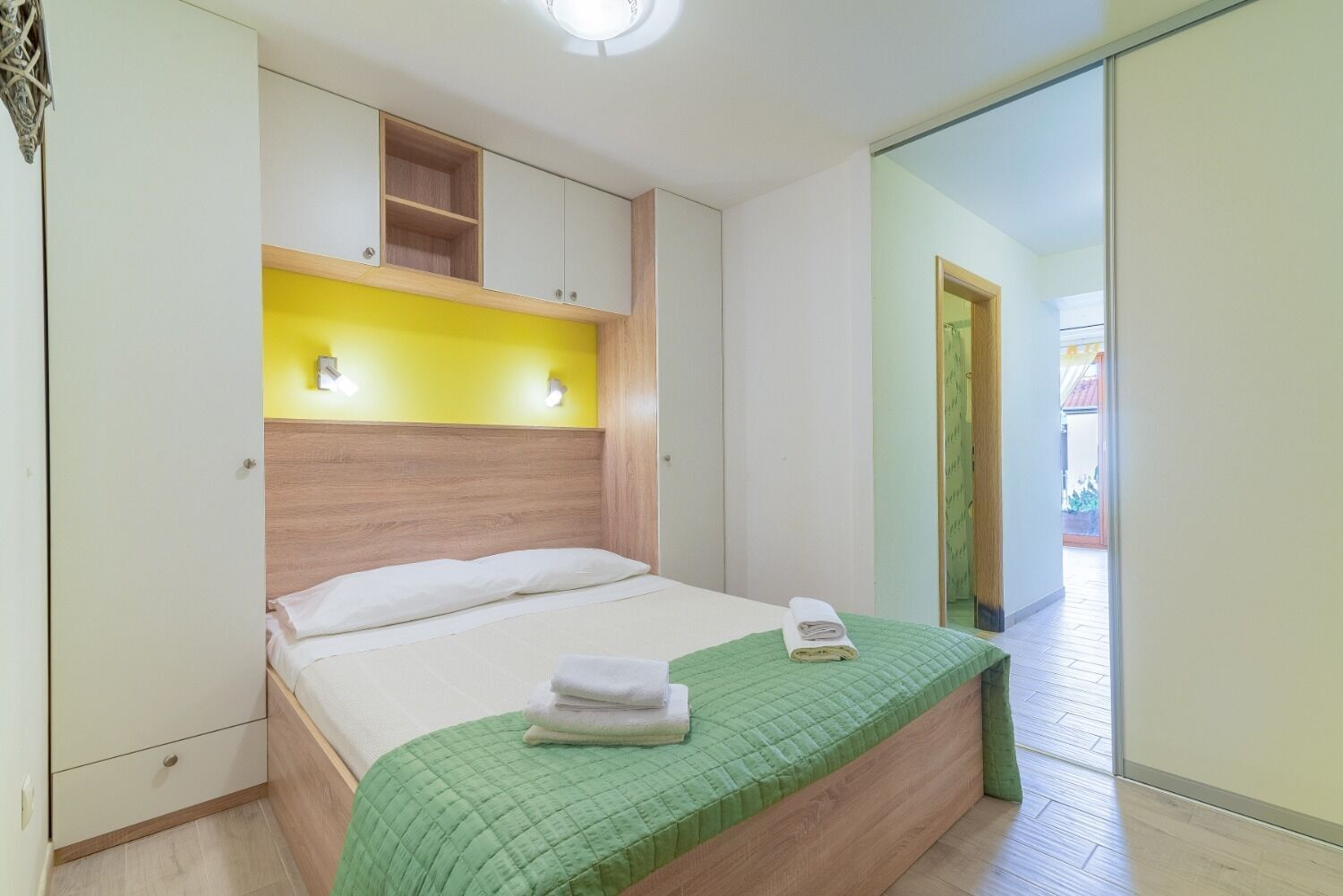 A bedroom with a bed, a green quilted bedspread, a wooden headboard, a yellow wall, and a sliding glass door leading to a balcony.