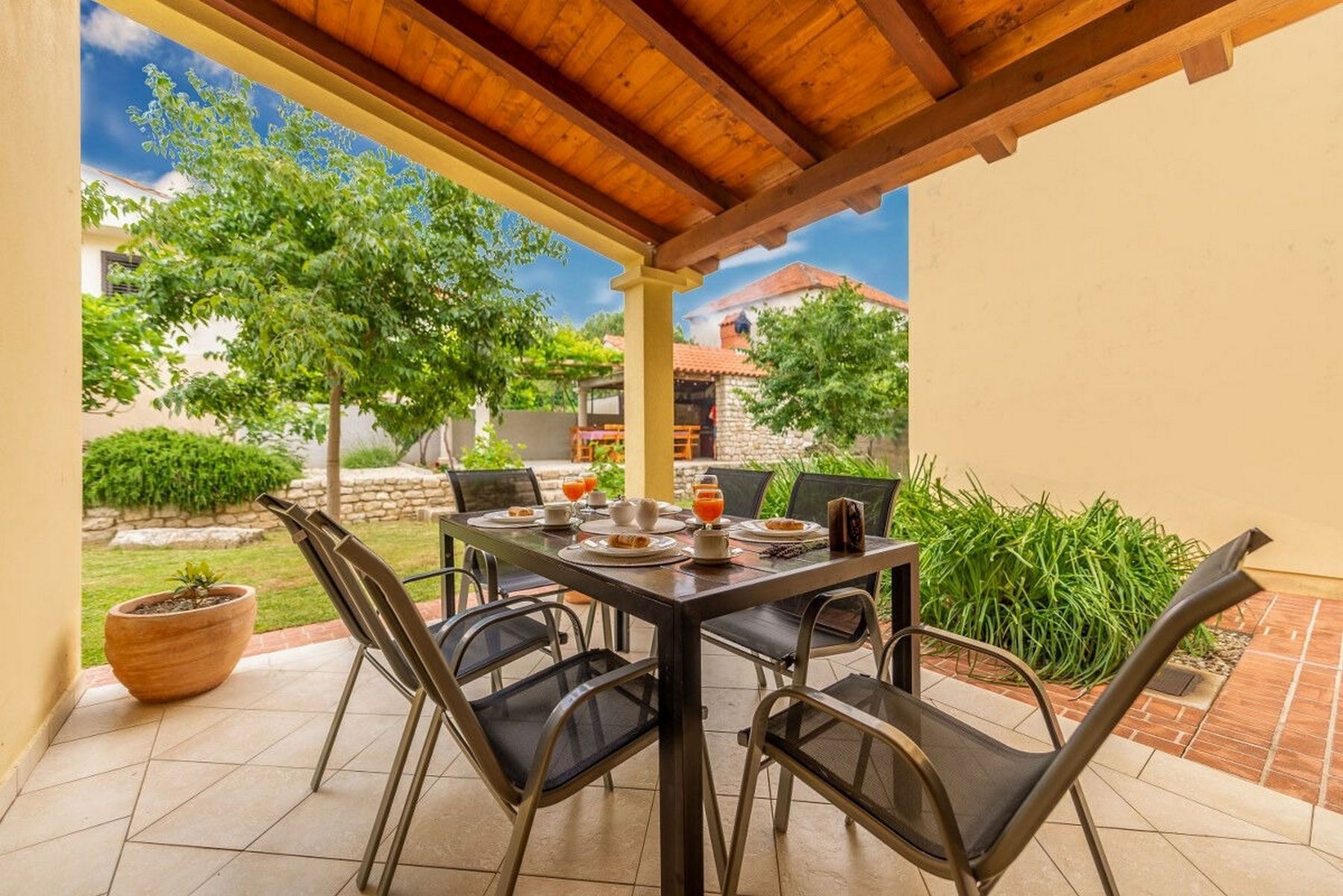 A covered patio with a table set for breakfast, surrounded by chairs, with a view of a garden and buildings in the background.