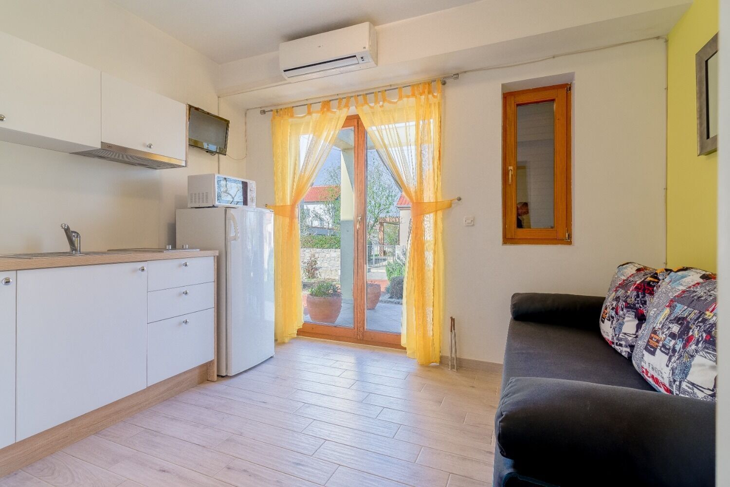 A compact kitchen with a refrigerator, microwave, and a small window. A sofa and a mirror are visible in the living area.