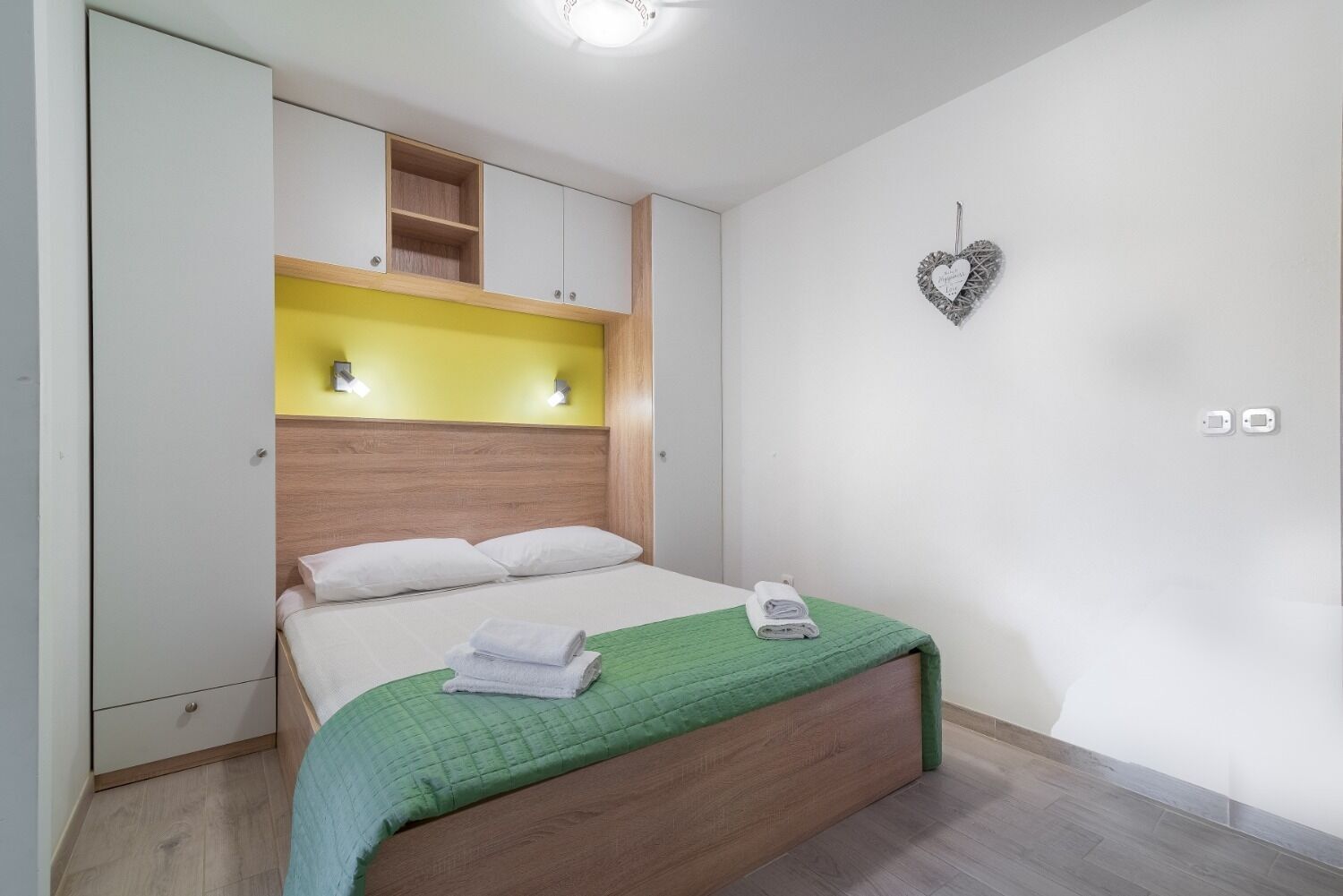 Un dormitorio con cama, cabecera de madera, colcha verde y decoración con forma de corazón en la pared.