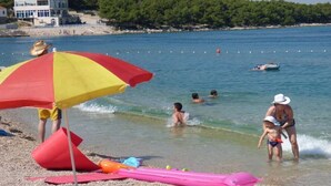 A beach with people swimming and playing, a large umbrella, and a building in the background. - Apartments Dragan (Tisno)