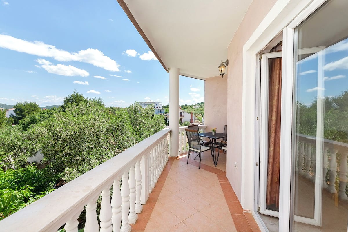 A balcony with a table and chairs, a white balustrade, and a view of greenery and buildings.