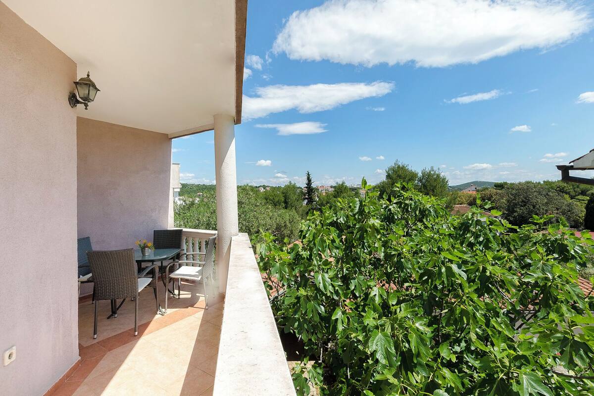 A balcony with a table and chairs, a potted plant, and a view of greenery and distant buildings.