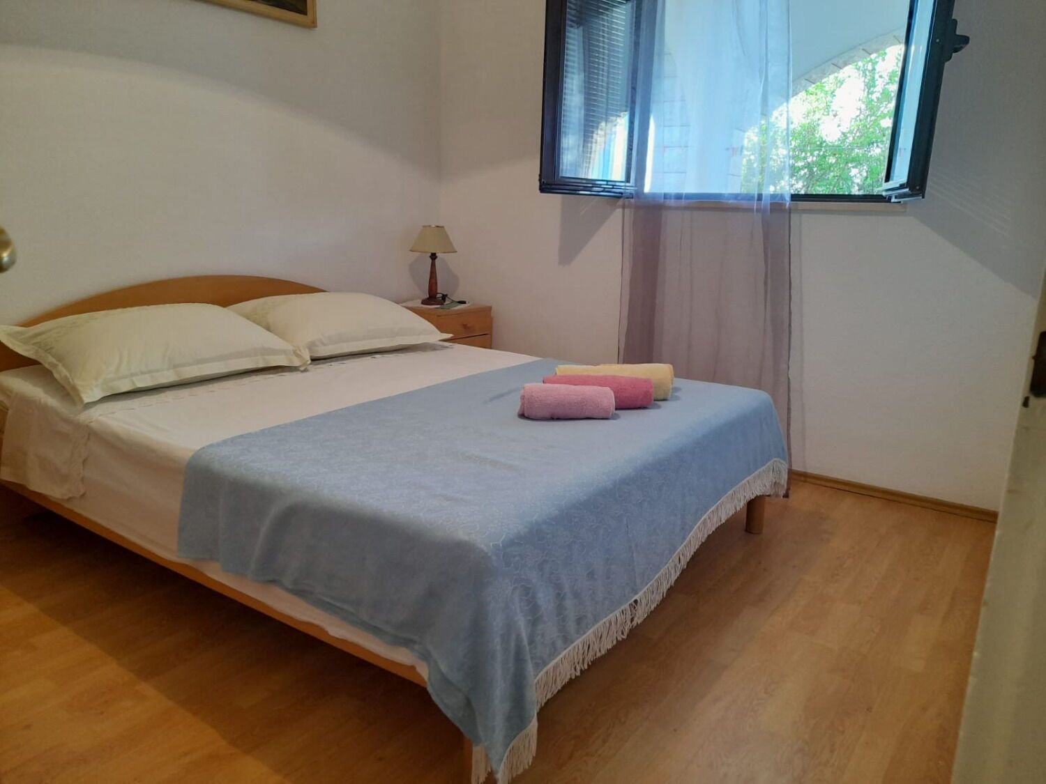 A neatly made bed with a blue bedspread and pillows, a bedside table with a lamp, and a window with curtains.