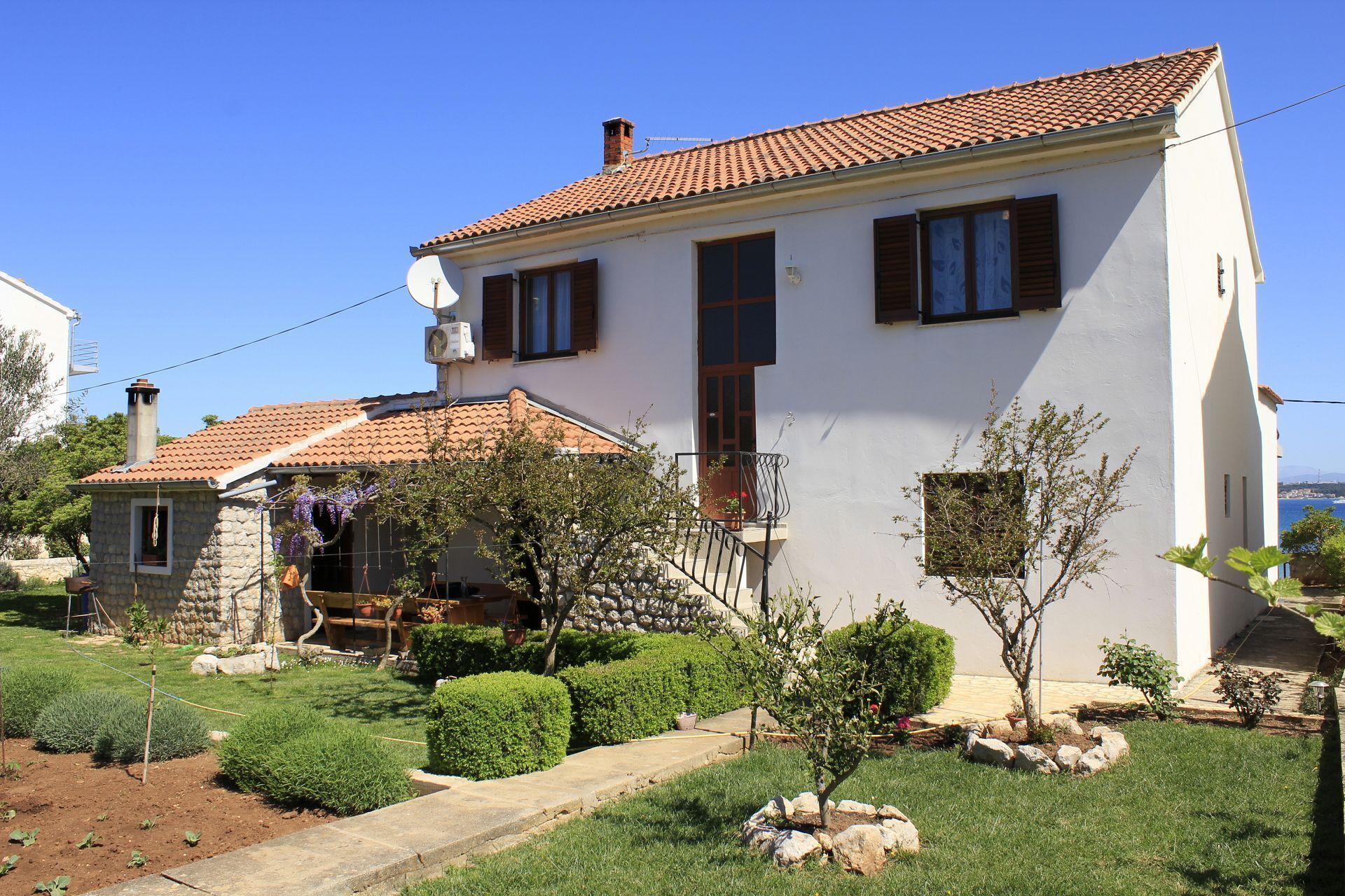 Una casa de dues plantes amb teulada de rajola, una antena parabòlica i un petit jardí amb arbustos i un arbre.