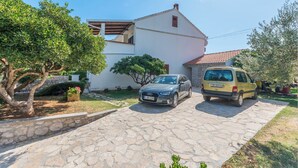 Una casa de dos pisos con camino de entrada de piedra, coches estacionados y follaje circundante.