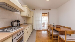 Cocina compacta con estufa a gas, horno y armarios blancos. Se observa una mesa de comedor con sillas al fondo.