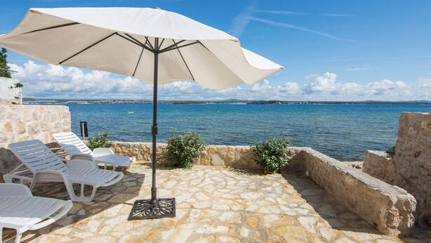 Un patio de piedra con sillones blancos y una sombrilla grande, con vistas a una masa de agua y un horizonte urbano distante.