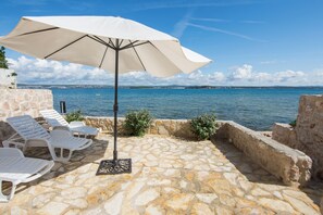 Une terrasse en pierre avec des chaises longues blanches et un grand parasol, offrant une vue sur une étendue d’eau et, au loin, sur le paysage urbain.