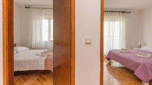 Un dormitorio con piso de madera, dos camas con ropa de cama de colores diferentes, una mesita de noche y un ventanal con cortinas traslúcidas.