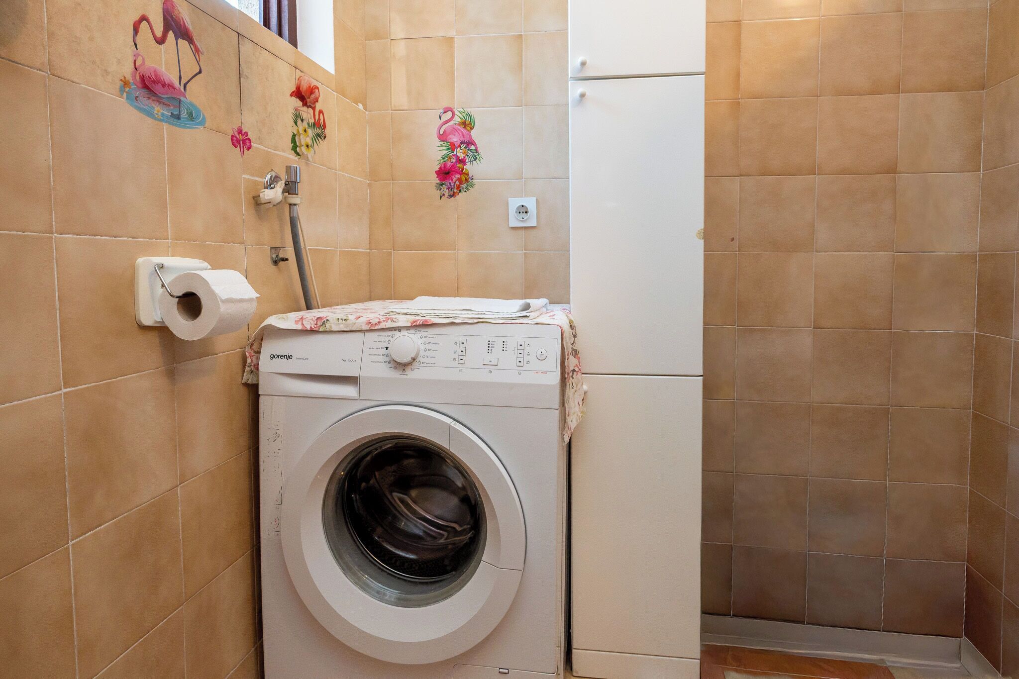 Un espace buanderie compact comprenant un lave-linge, un distributeur de papier toilette et des carreaux décoratifs sur les murs.