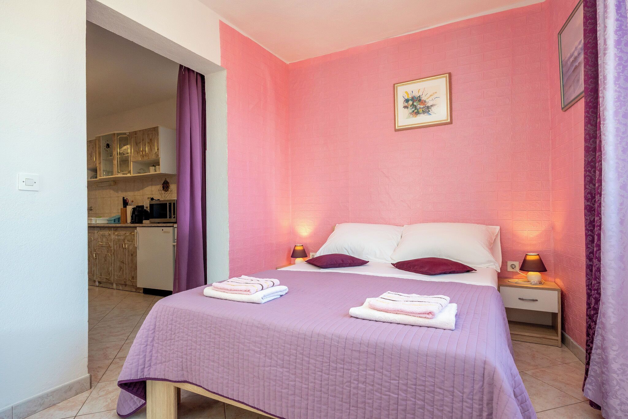 Quarto com cama, almofadas, mesa de cabeceira, abajur e banheiro ao fundo.