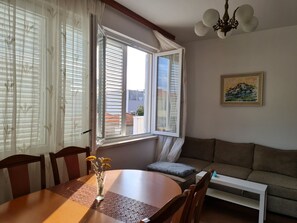 A dining area with a wooden table, chairs, and a vase with dried flowers. There is a window with blinds, a sofa, and a painting on the wall. - Apartments Dami (Orebic)