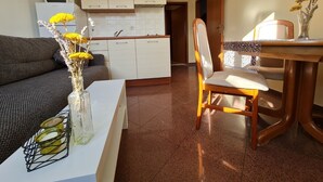 En stue med en sofa, et spisebord med en stol, en vase med blomster og et køkken i baggrunden.